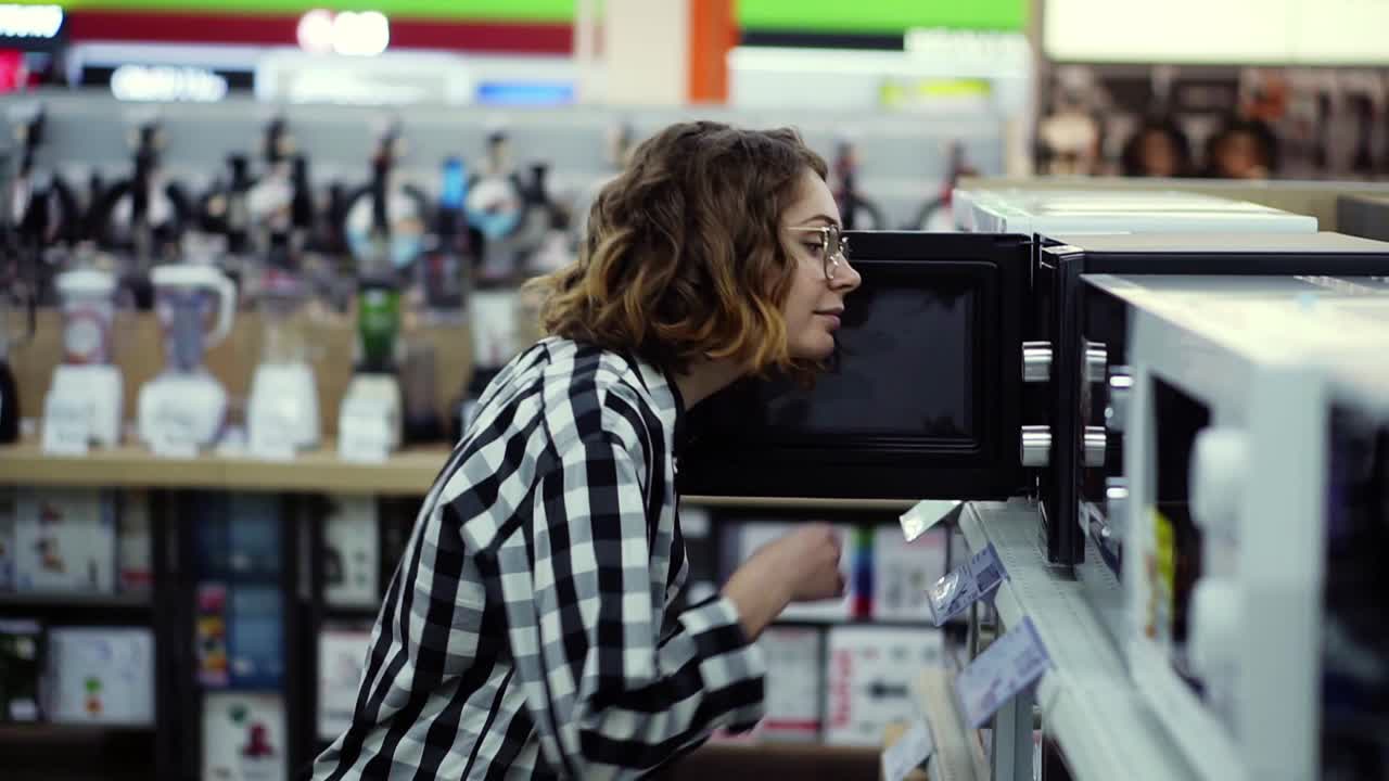 Side view of a young curly haired woman in plaid shirt chooses a microwave oven in a consumer electronics supermarket. Opens the