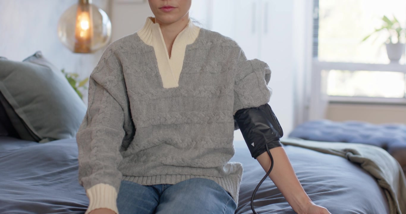 Donna afroamericana seduta sul letto con un maglione, con un bracciale collegato a un monitor che registra la pressione sanguigna.