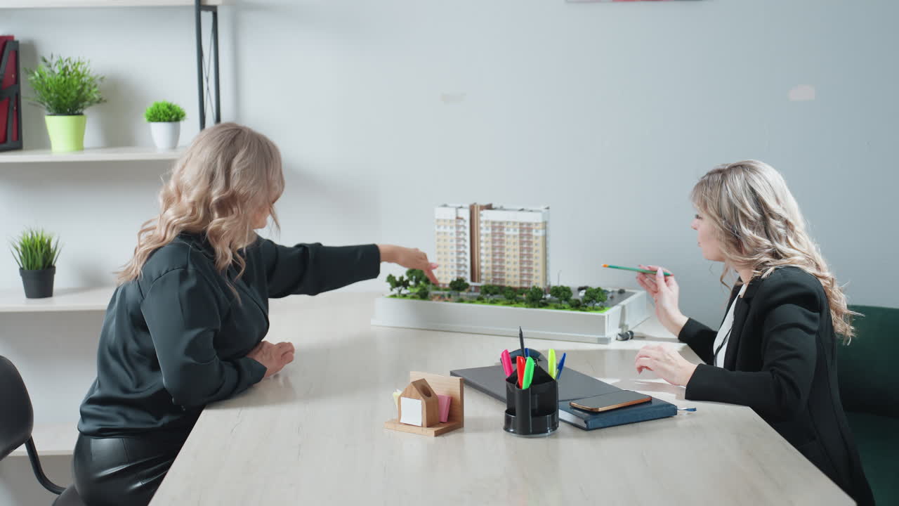 Realtor discussing residential building model with client during office meeting, using pencil to explain details of property project, both women engaged in professional conversation