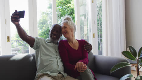 Una-Pareja-De-Ancianos-De-Raza-Mixta-Tomando-Un-Selfie-Con-Un-Teléfono-Inteligente-En-El-Salón-De-Casa