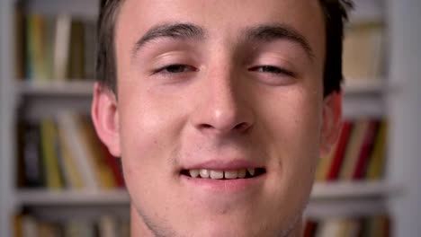 face of young attractive businessman is watching at camera, smiling, bookshelf on background, blurred background