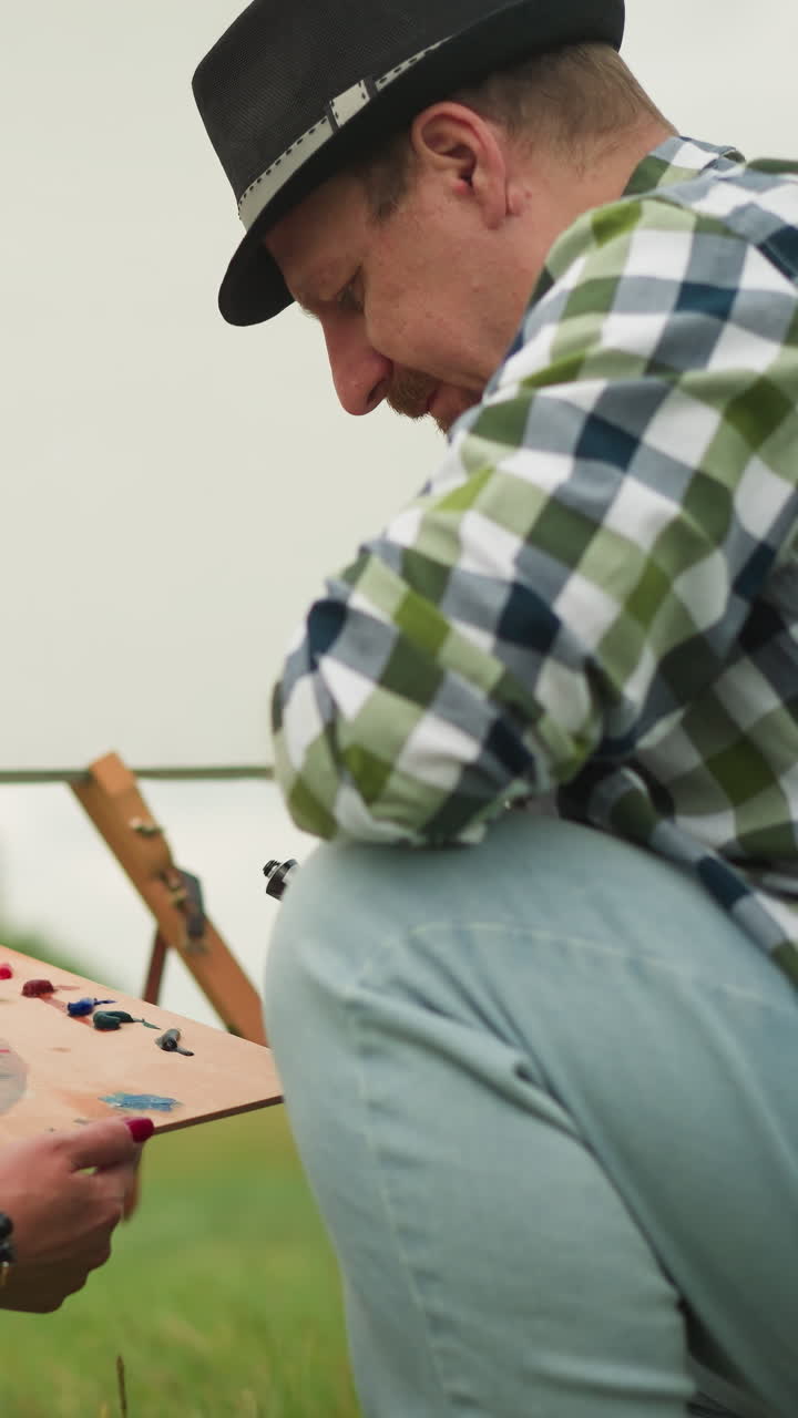 un hombre y una mujer se inclinan en un campo cubierto de hierba, preparando una sesión de pintura al aire libre. la mujer con un vestido blanco sostiene una paleta de madera, mientras que el hombre con una camisa a cuadros exprime cuidadosamente la pintura