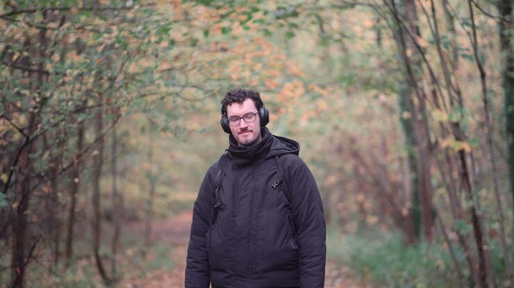 Cheerful young German gentleman with glasses and beard, stands amidst the hues of an autumnal European mixed forest, grinning warmly and offering a friendly greeting