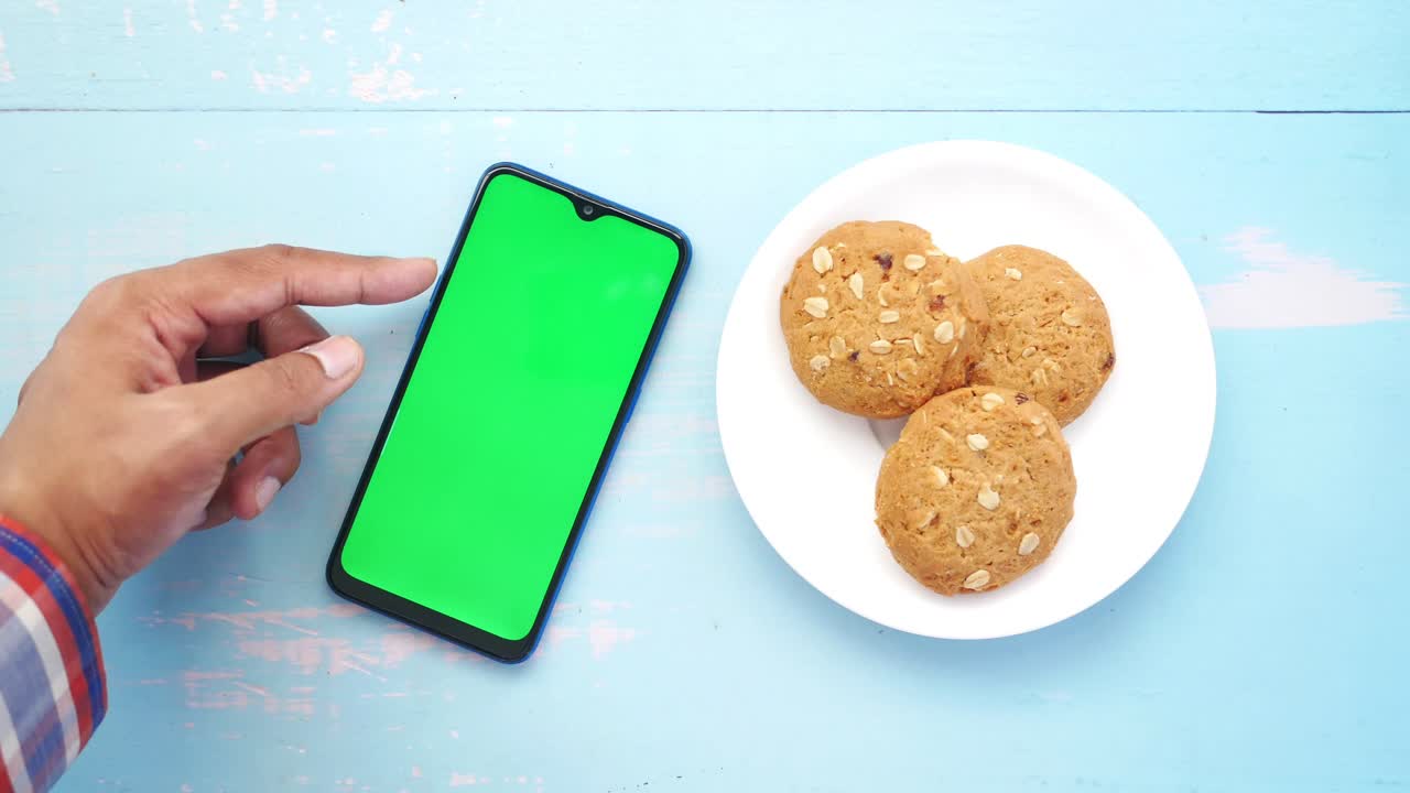 mano apuntando a un teléfono inteligente de pantalla verde con galletas en un plato.