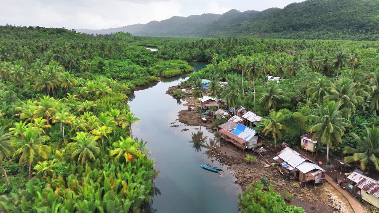 A tranquil river winds through dense mangroves and palm forests beside a remote village in Mataob Maasin, Siargao Islands, Philippines, nestled against green hills under a cloudy sky