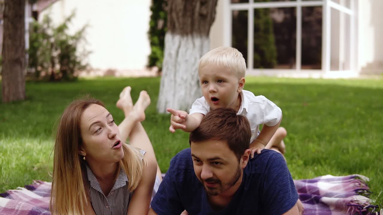 el niño está jugando al aire libre con sus padres. el padre y la madre descansan en el suelo, hacen picnic. el hijo está en la espalda del padre, apuntando con el dedo en alguna parte. ropa informal. día soleado, día de fiesta