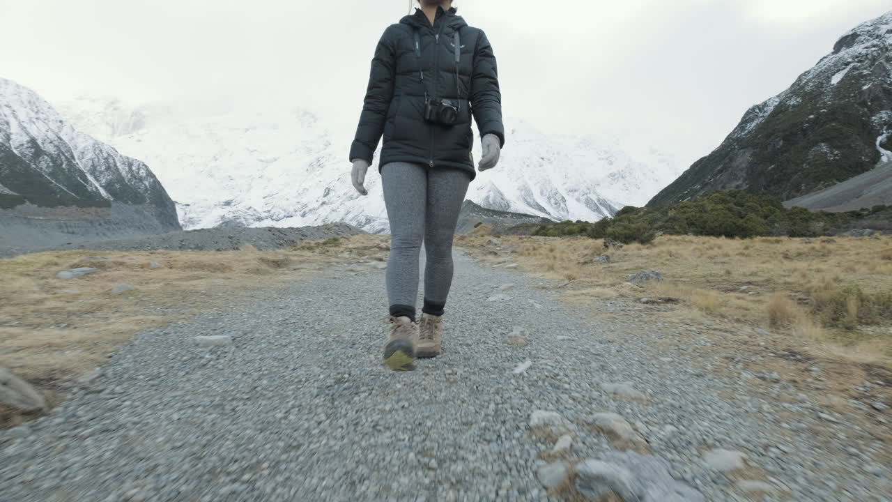 Et følgeskudd fra lav til høy vinkel av en kvinne som vandrer mellom snøkledde fjell i New Zealand en kald vintermorgen.