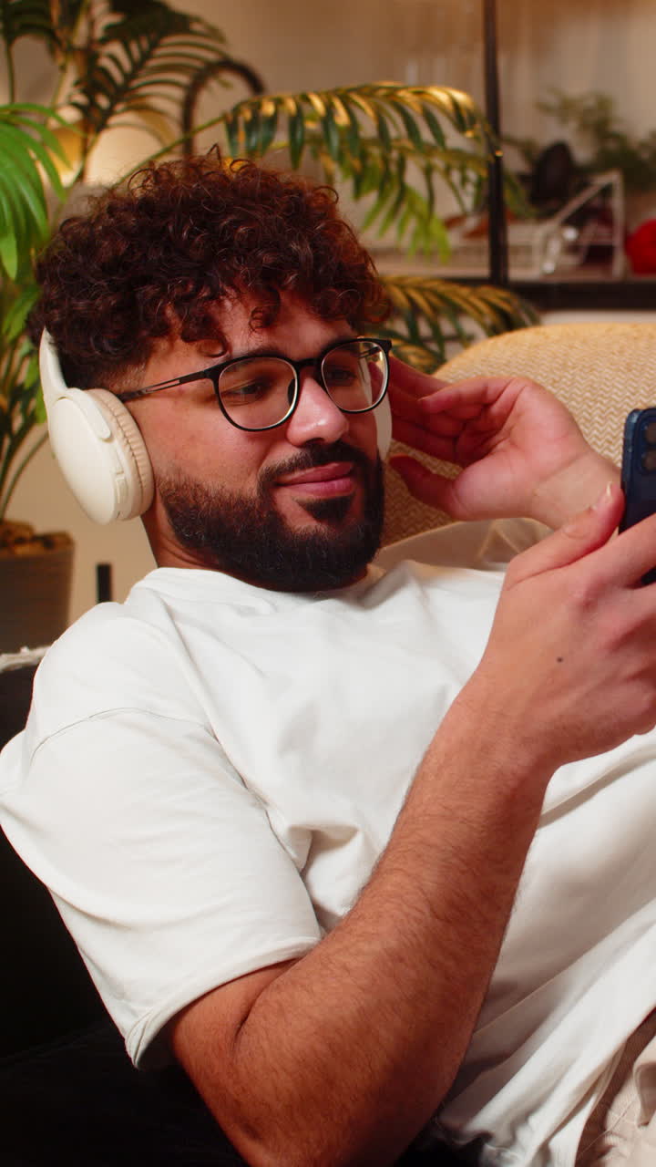 Arabisk mann med trådløse hodetelefoner lytter til musikktekster på smarttelefonen om natten innendørs i avslappet modus
