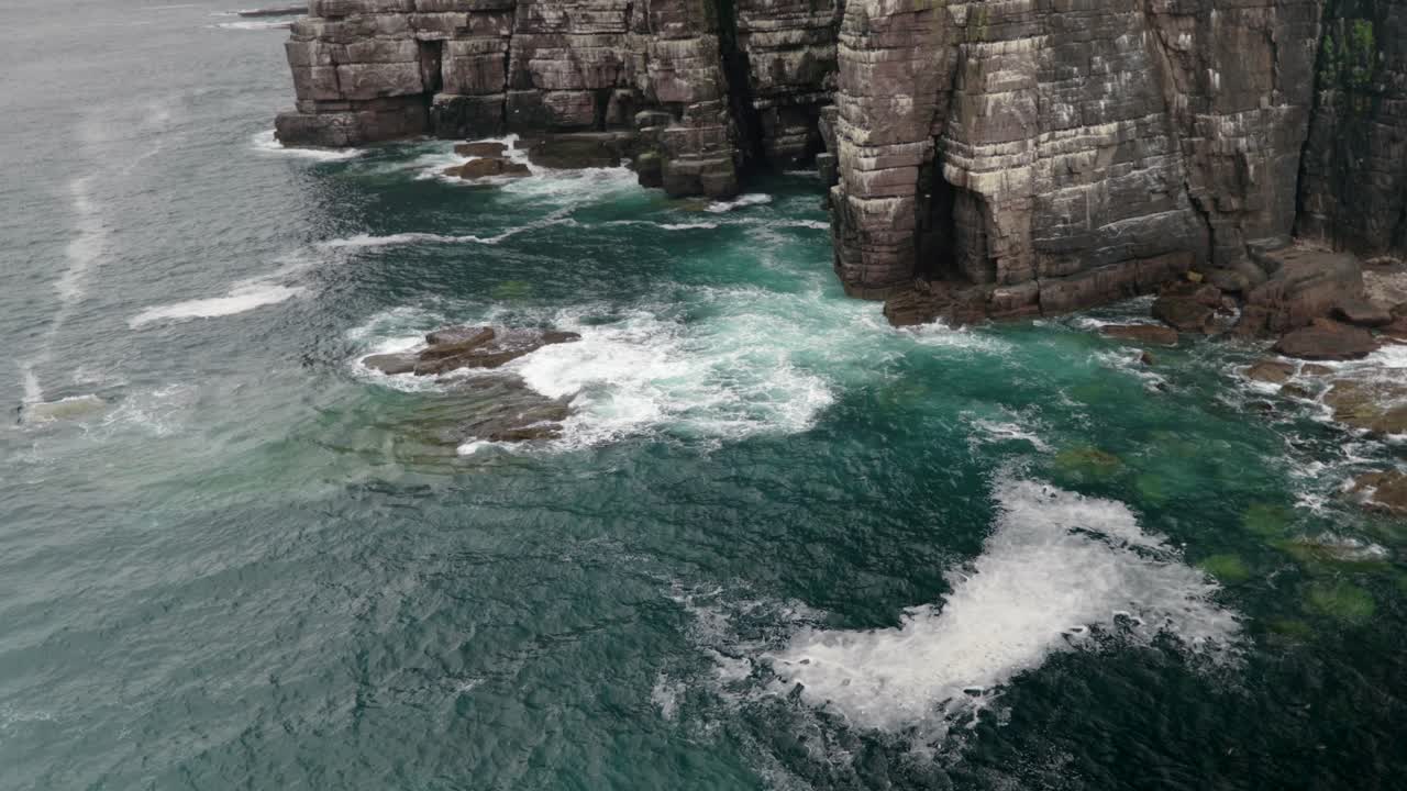Waves gently crash over rocks and against a tall and imposing sea cliff in the deep teal coloured ocean while seabirds fly around the cliffs of a seabird colony of guillemots on Handa Island, Scotland