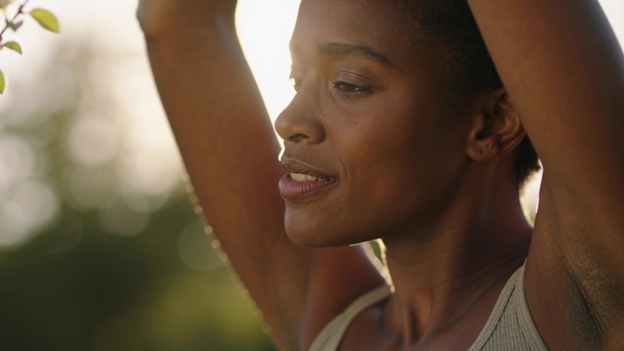 Smiling lowering gaze woman in ribbed tank clasping hands lifting arms stretching to branch at park