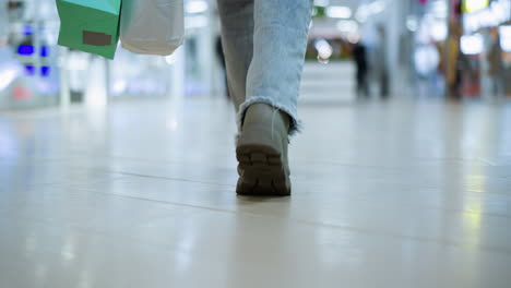 vista de la pierna trasera de alguien con zapatos caros y vaqueros caminando en un centro comercial bien iluminado con una bolsa de compras, figuras borrosas moviéndose en el fondo