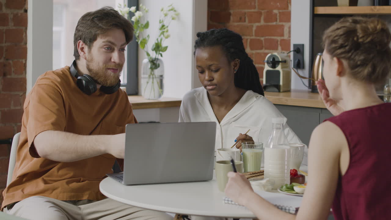 Boy Using Laptop Computer To Explain Something To His Two Female Roommates While Sitting Together In The Kitchen During Breakfast