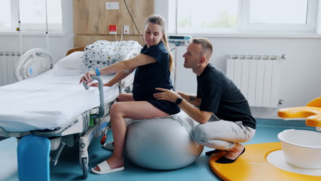 pregnant woman sitting on a fitball in the hospital ward. her husband sitting behind massaging wife's back.