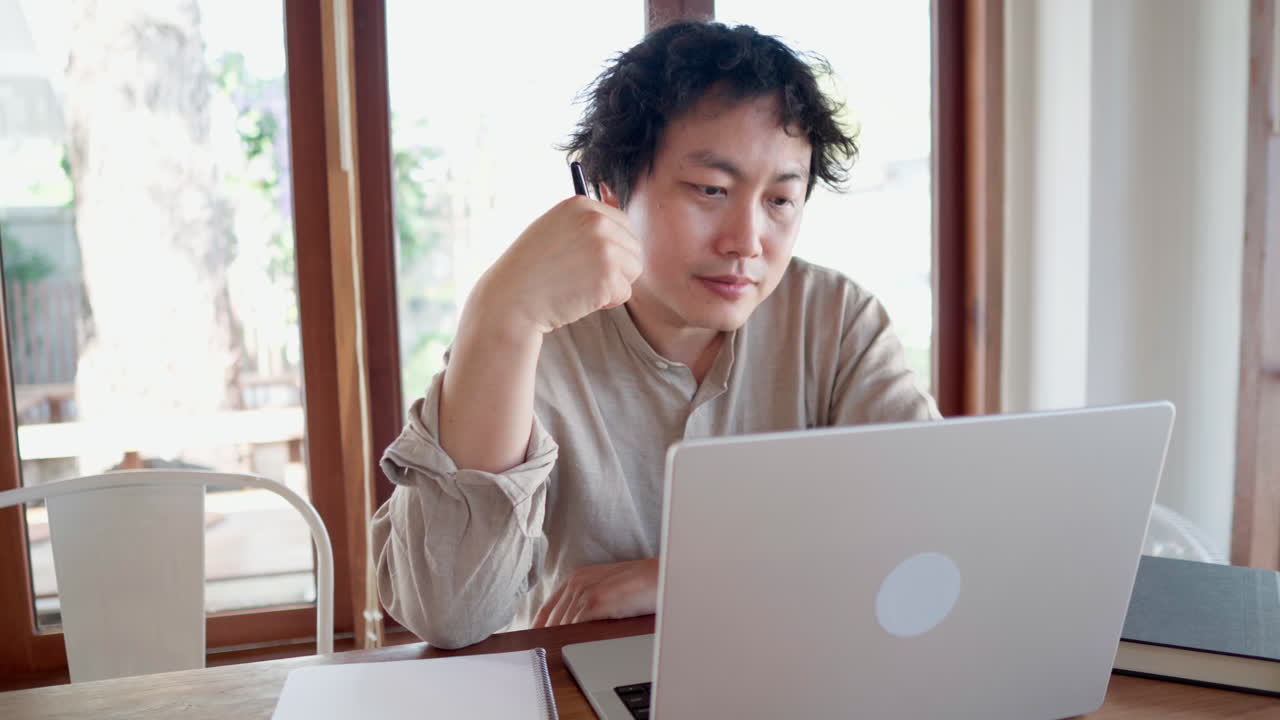 집중된 심각한 아시아인 노트북에서 온라인 학습 과정을 보고 메모를 취합니다.