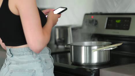 mujer caucásica parada frente a la estufa, en su teléfono, mientras el vapor sale de la olla de pasta para cocinar en la estufa