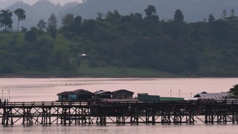 Mon-Bridge-and-a-longboat-followed-speeding-behind-the-structure-while-people-are-walking-on-the-bridge,-silhouetting-as-it-was-getting-dark,-in-slow-motion