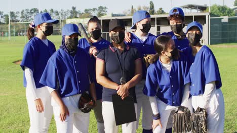 group portrait of diverse team of female baseball players and coach in face masks on sunny pitch