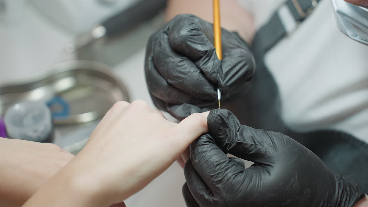 Nail technician in black glove carefully uses tiny brush to apply polish on customer fingernail during detailed manicure session under bright light with tools and tray visible on salon table