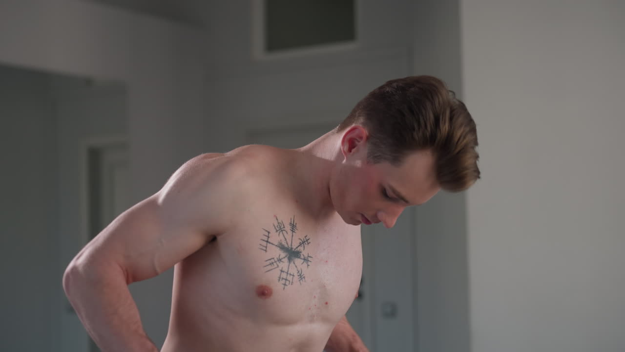 shirtless young man with chest tattoo intensely focused during workout session in minimalist room with closed white door and large wall mirror behind, arms engaged, body leaning forward