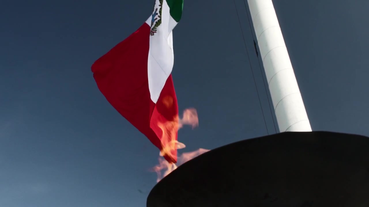 Primo piano di una grande bandiera messicana che sventola su un'alta asta contro un cielo azzurro limpido, con il tessuto rosso, bianco e verde in movimento.