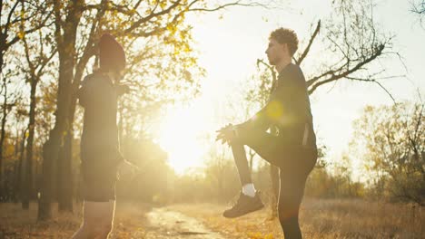 a man with curly hair in a black sports uniform together with his friend in a red cap and black shorts are warming up before their jogging in the autumn park at sunrise on a sunny morning in autumn