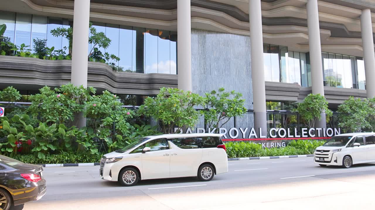 Camera pans down lush, multi-story building facade as cars drive past in bright daylight
