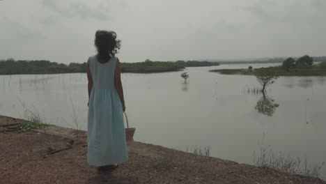 Static-slow-motion-shot-of-a-pretty-young-woman-dressed-in-light-blue-dress,-black-hair-and-a-wood-basket-in-her-hand-looking-in-front-of-a-beautiful-lake-with-sea-islands-during-a-windy-weather