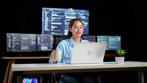 asian female programmer drinking coffee while writing code by a laptop using multiple monitors showing database on terminal window desktops in the office