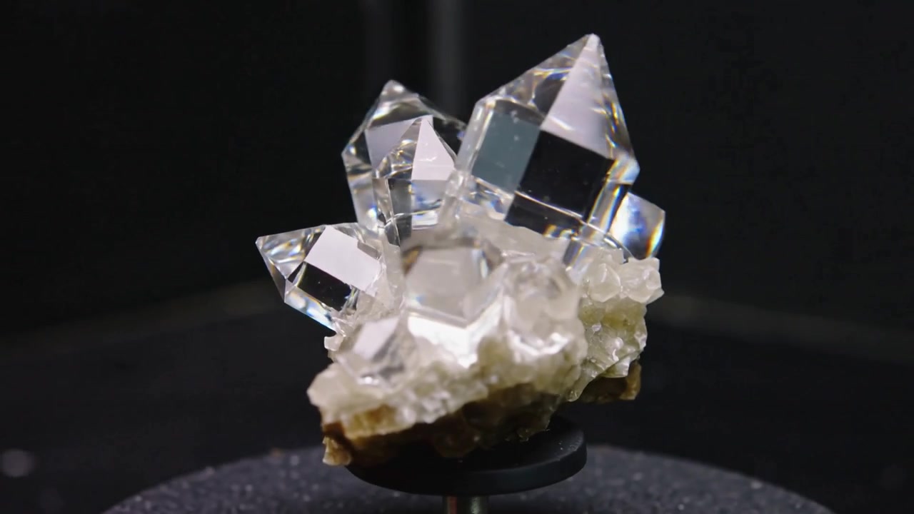 Bout du doigt effleurant un amas de quartz transparent tournant sur un support noir pour révéler des facettes prismatiques éclatantes
