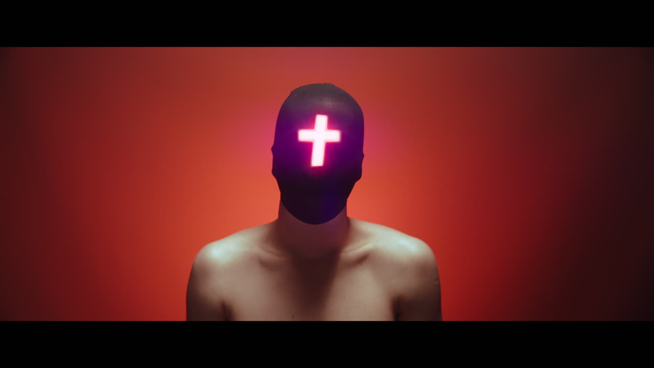 Masked man with cross of light glowing on his face nods gently