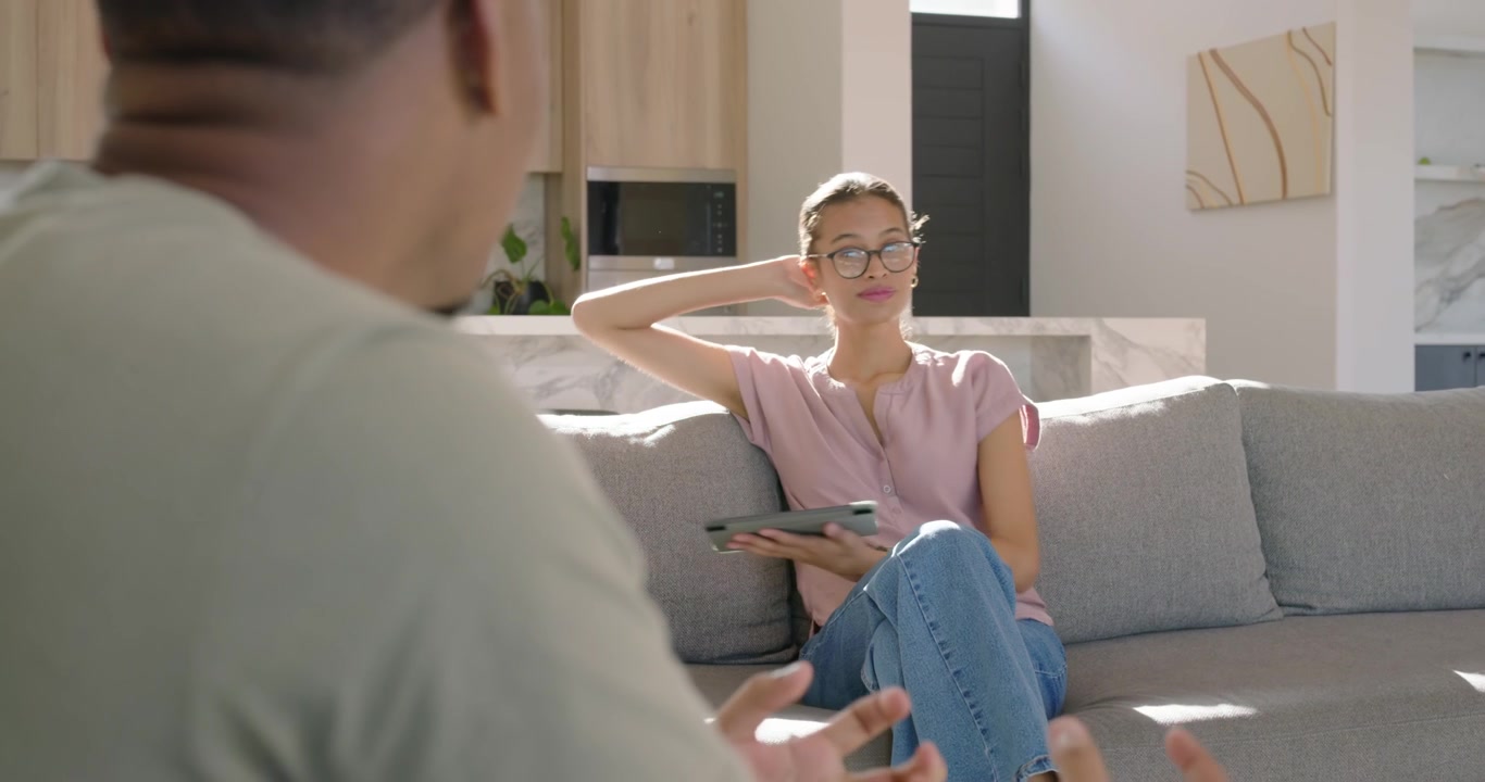 Par sidder på grå sofa, mand taler og gestikulerer, kvinde sænker tablet og svarer.