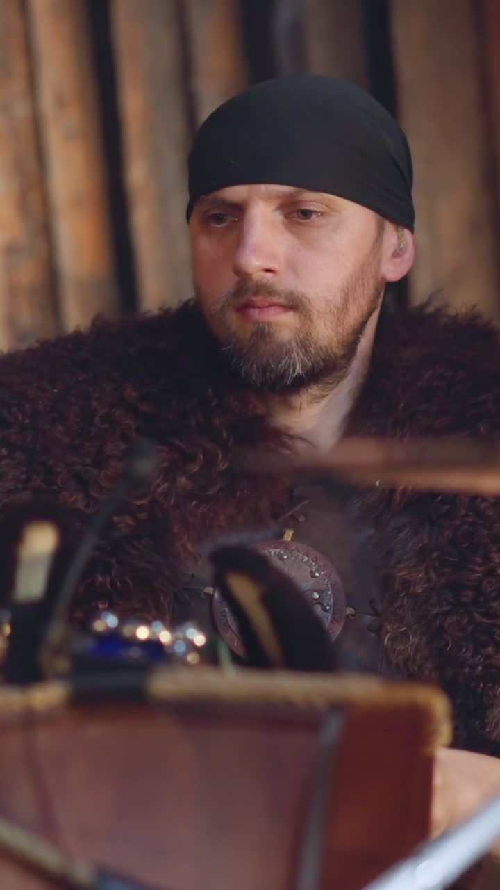 Bearded man plays drums sitting near wall of rough logs slow motion. Musician with fur on shoulders performs with old instruments. Concert of folk group