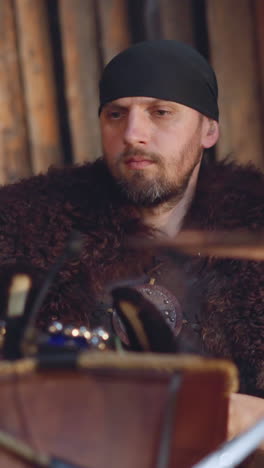 bearded man plays drums sitting near wall of rough logs slow motion. musician with fur on shoulders performs with old instruments. concert of folk group