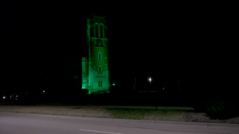 Beaumont-Tower-on-the-campus-of-Michigan-State-University-lit-up-at-night-in-green-in-honor-of-the-victims-of-the-February,-2023-mass-shooting-with-wide-shot-with-cars-driving-by