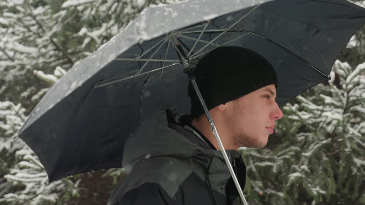 Profile white man walking under umbrella, purposeful stride along snowdusted trail, black beanie and insulated jacket, gloved hand gripping umbrella, side profile framed by evergreen branches, cold