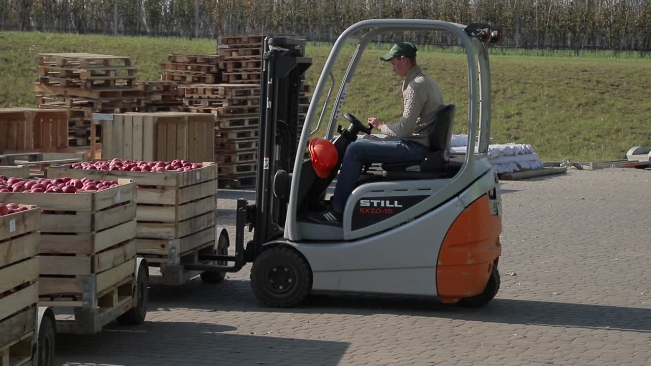 A man on a new forklift truck pulls up quickly to the pallet with red apples, lifts it and takes it to a warehouse. Close-up.