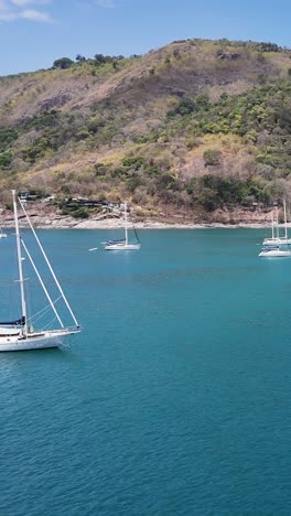 drone footage captures luxury yachts sailing near phuket's coastline, showcasing clear waters and lush hills under bright daylight
