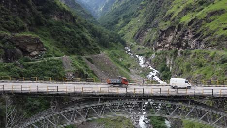 Eine-Drohnenaufnahme-Von-Nutzfahrzeugen,-Die-Auf-Einer-Hängebrücke-In-Einem-Flusstal-In-Der-Abgelegenen-Autobahn-Von-Himachal-Pradesh-Am-Fuße-Des-Indischen-Himalaya-Fahren