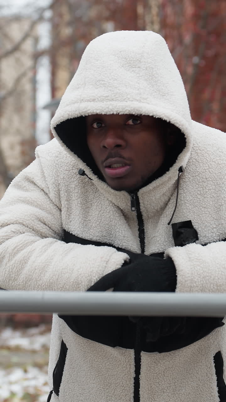 Music artist in white hoodie and gloves rests on iron bar outdoors, cold air visible from mouth, background features urban buildings, snowy ground, and trees