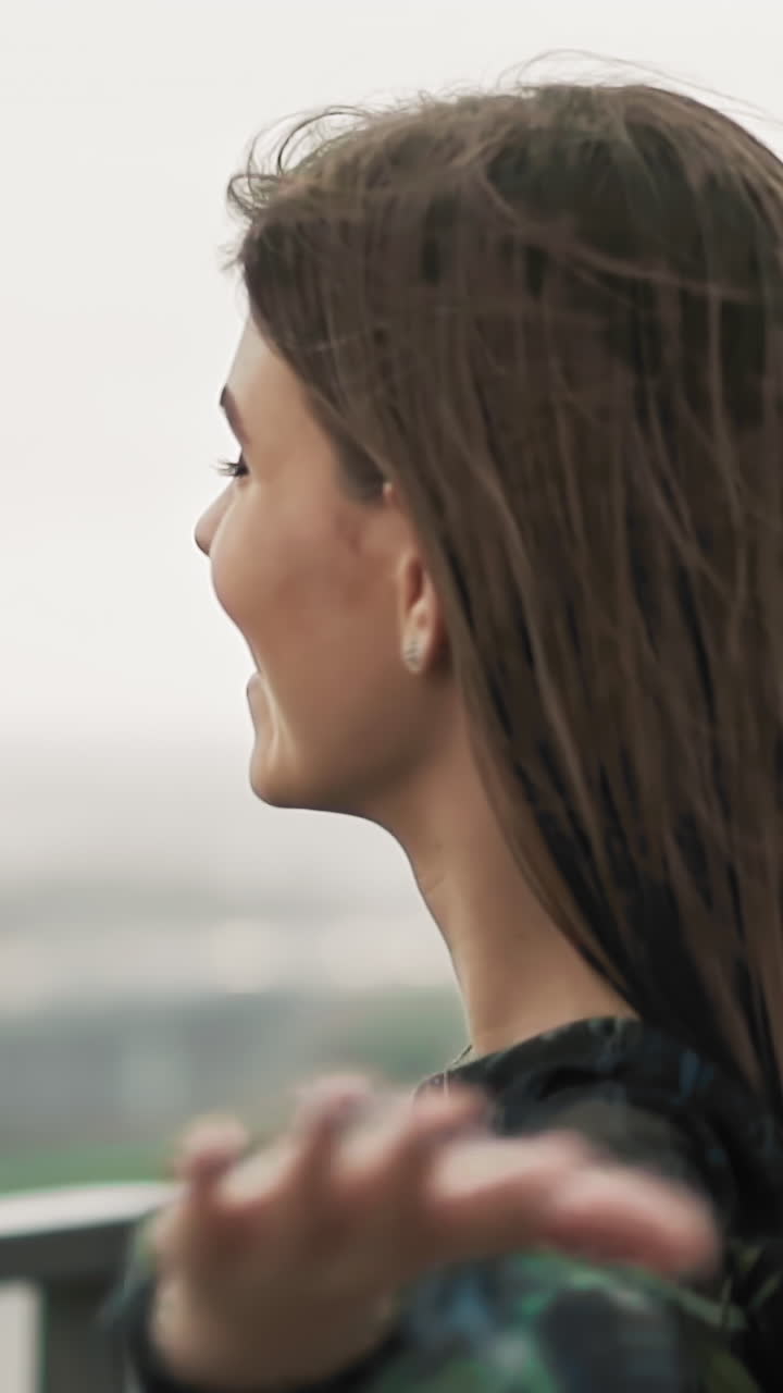Joyful young woman in elegant dress with large black umbrella turns around standing in city park under light spring rain slow motion close view