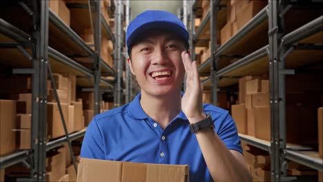 primer plano de un mensajero masculino asiático en uniforme azul sonriendo y hablando con la cámara mientras entrega un cartón en el almacén