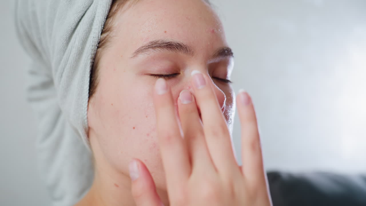 Closeup of young girl with towel wrapped around head applying skincare cream on face with finger, eyes closed, practicing facial self-care, treating acne-prone skin, focused on beauty