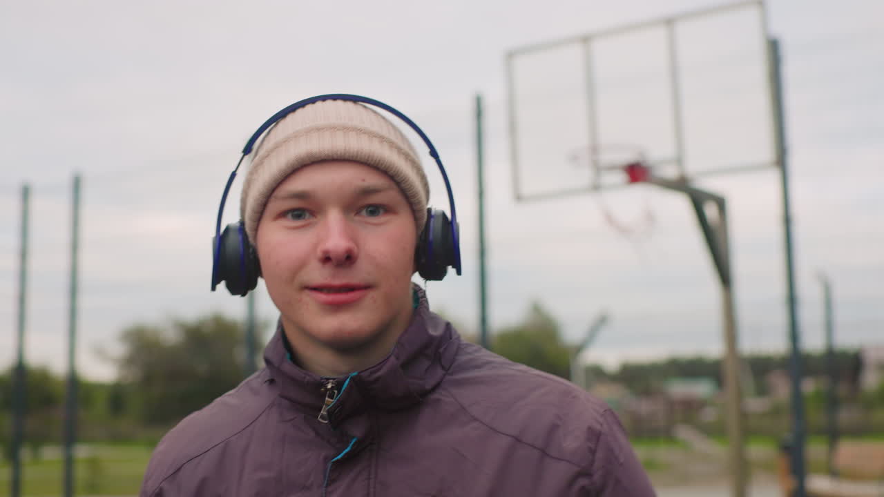 Homem branco jovem de fones de ouvido em uma quadra de basquete. Jogador casual faz uma pausa entre os exercícios, usando gorro e jaqueta. Céu nublado, aro de metal e cerca ao fundo. Mãos seguram a bola de basquete, sorriso relaxado, ouvindo.