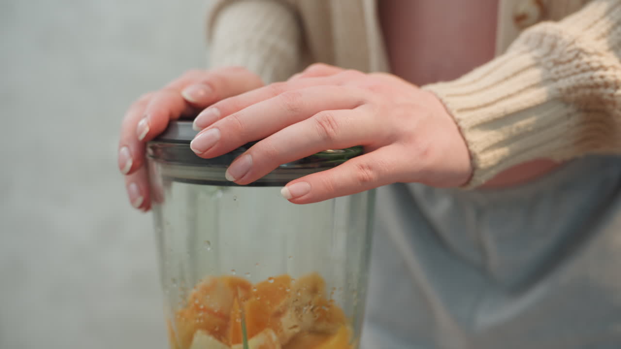 Mujer caucásica embarazada preparando una licuadora de frutas, primer plano de manos añadiendo trozos de naranja y aguacate en un frasco de vidrio, cárdigan suave, vientre expuesto, suave luz matutina, cocina hogareña acogedora, prenatal.
