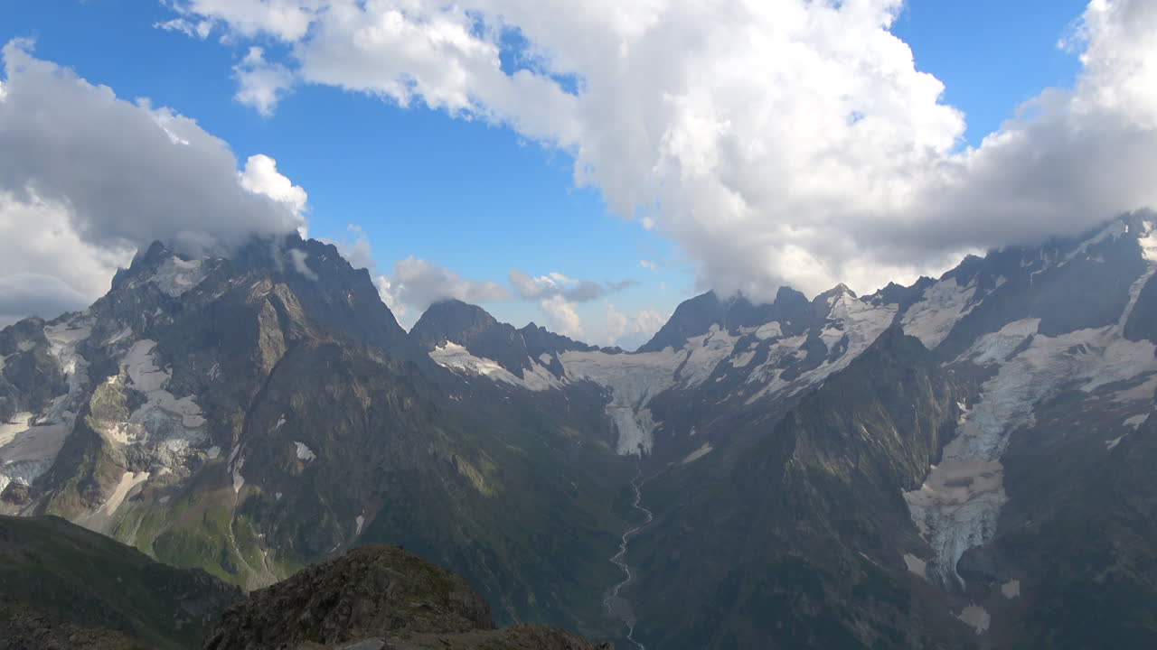 Pemandangan panorama luas Pegunungan Kaukasus yang terjal dengan padang salju dan gletser di bawah awan dramatis dekat Dombay, Rusia, ideal untuk latar belakang perjalanan dan alam.