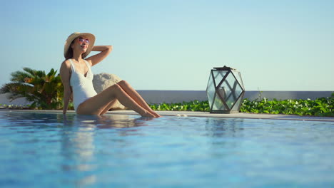 Mujer-Asiática-Sexy-En-Monokini-Blanco-Se-Sienta-En-El-Borde-De-La-Piscina,-Disfruta-Del-Sol