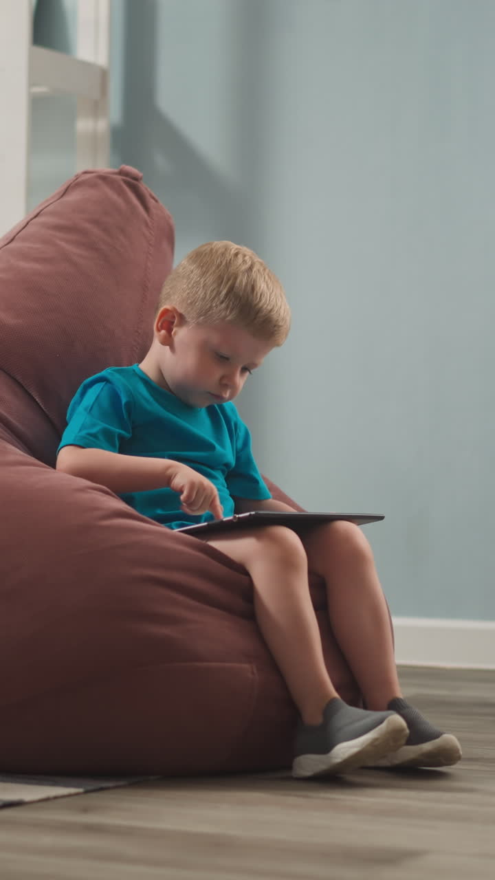 Adorable toddler boy plays video game on tablet computer sitting in bean chair at home. Development application for little children. Domestic lifestyle