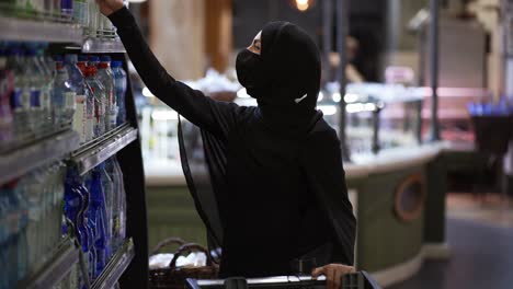 woman in hijab and protective mask doing shopping, takes bottle of water from the shelf