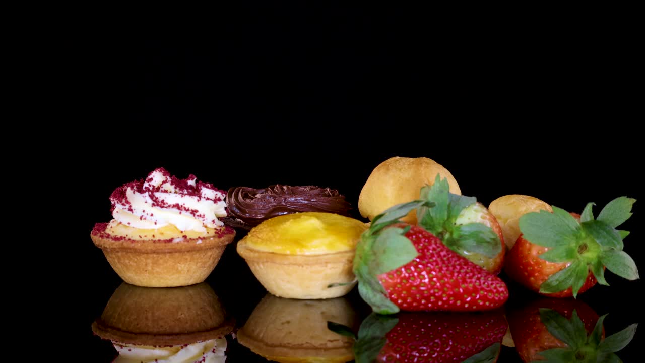 A selection of desserts including cupcakes, tarts, cream puffs, and fresh strawberries rotates smoothly on a reflective black surface under bright studio lighting
