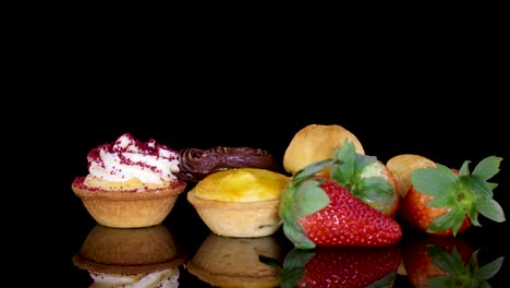 a selection of desserts including cupcakes, tarts, cream puffs, and fresh strawberries rotates smoothly on a reflective black surface under bright studio lighting